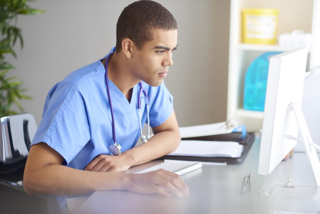 male nurse or doctor sits at his desk looking at his computer screen and writing in his pad . Sunlight pours across the desk from the window beside the desk. He is wearing scrubs .