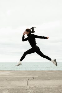 Woman in athletic wear jumps energetically by the ocean on a cloudy winter day.