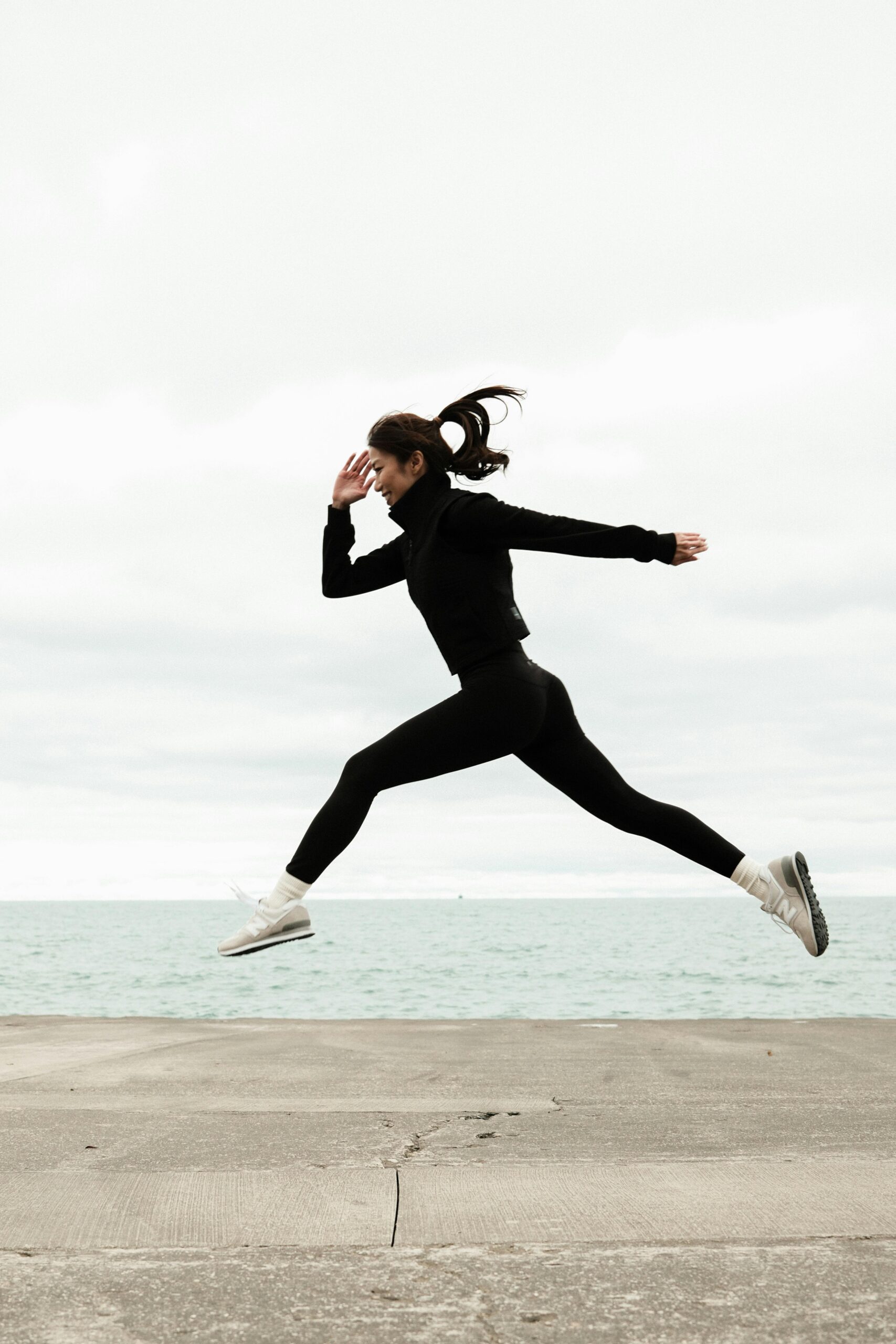 Woman in athletic wear jumps energetically by the ocean on a cloudy winter day.