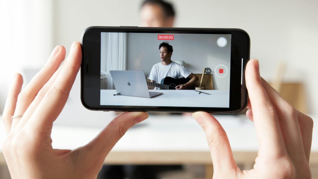 Man playing guitar while recording a vlog video using a smartphone.