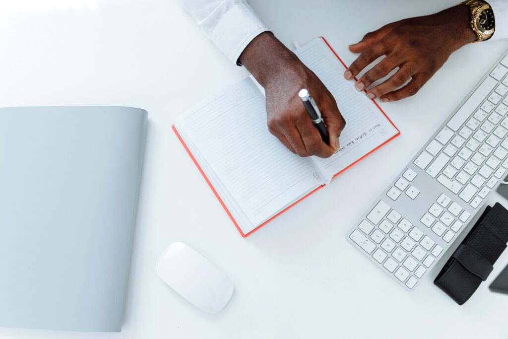 Businessman writing in a notebook on a white desk with keyboard and mouse.